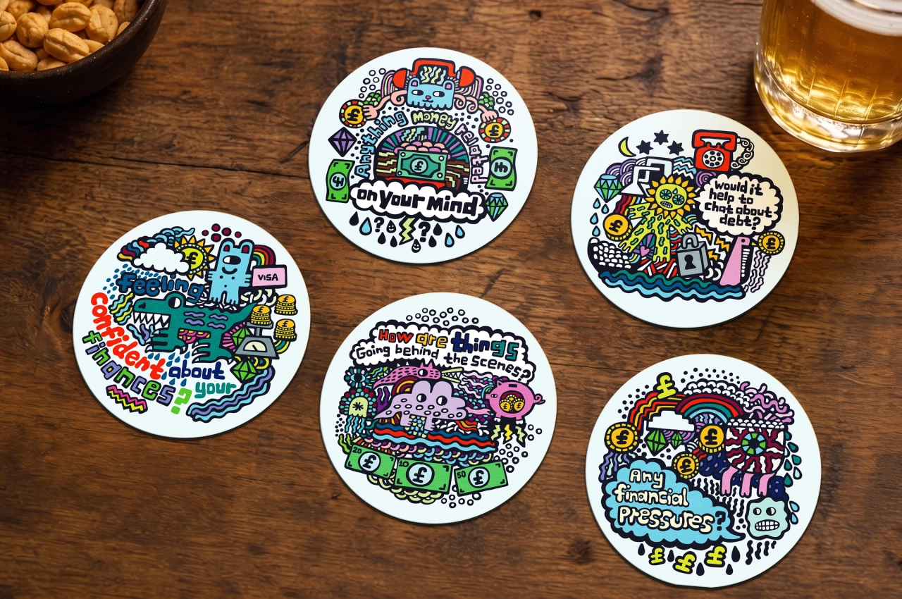 Five illustrated coasters with colourful doodle designs and text about debt conversations, arranged on a wooden table with snacks and a beer glass