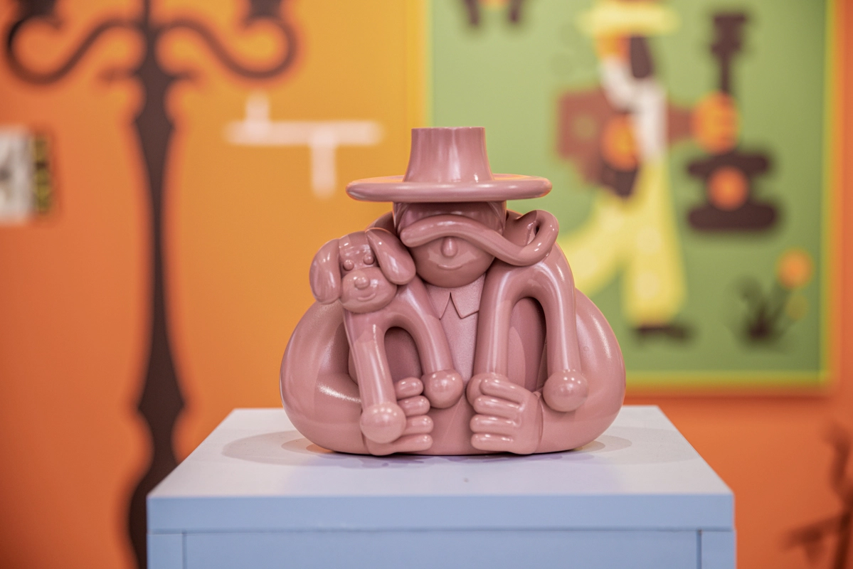 Terracotta-coloured ceramic sculpture of a rotund figure wearing a wide-brimmed hat, displayed on a light blue plinth against a colourful gallery wall