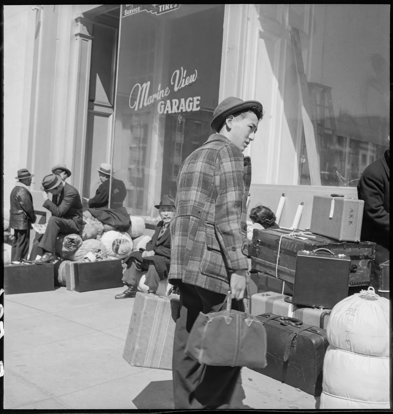 Man in plaid jacket carrying suitcase outside Alpine View Garage during Japanese American evacuation to Manzanar internment camp, 1942