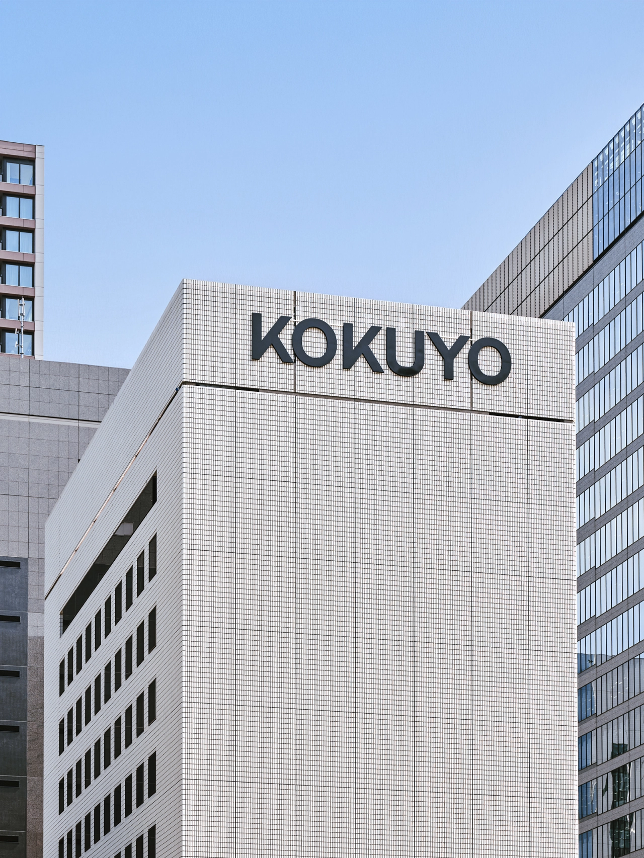 Kokuyo's modernist white office building with the company name displayed prominently in large black lettering on its facade, flanked by contemporary multi-storey buildings under clear blue sky