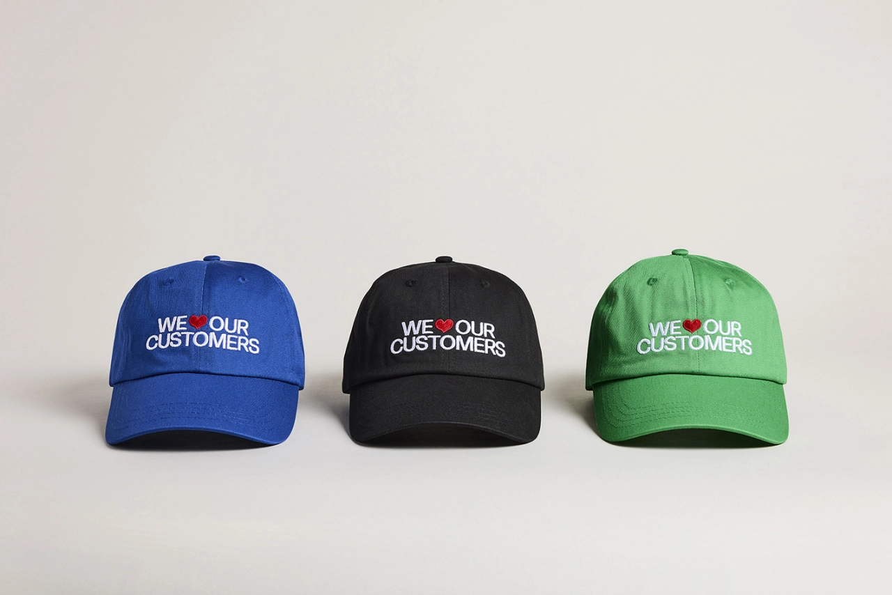 Three baseball caps in blue, black and green, each printed with "WE LOVE OUR CUSTOMERS" and a small heart logo