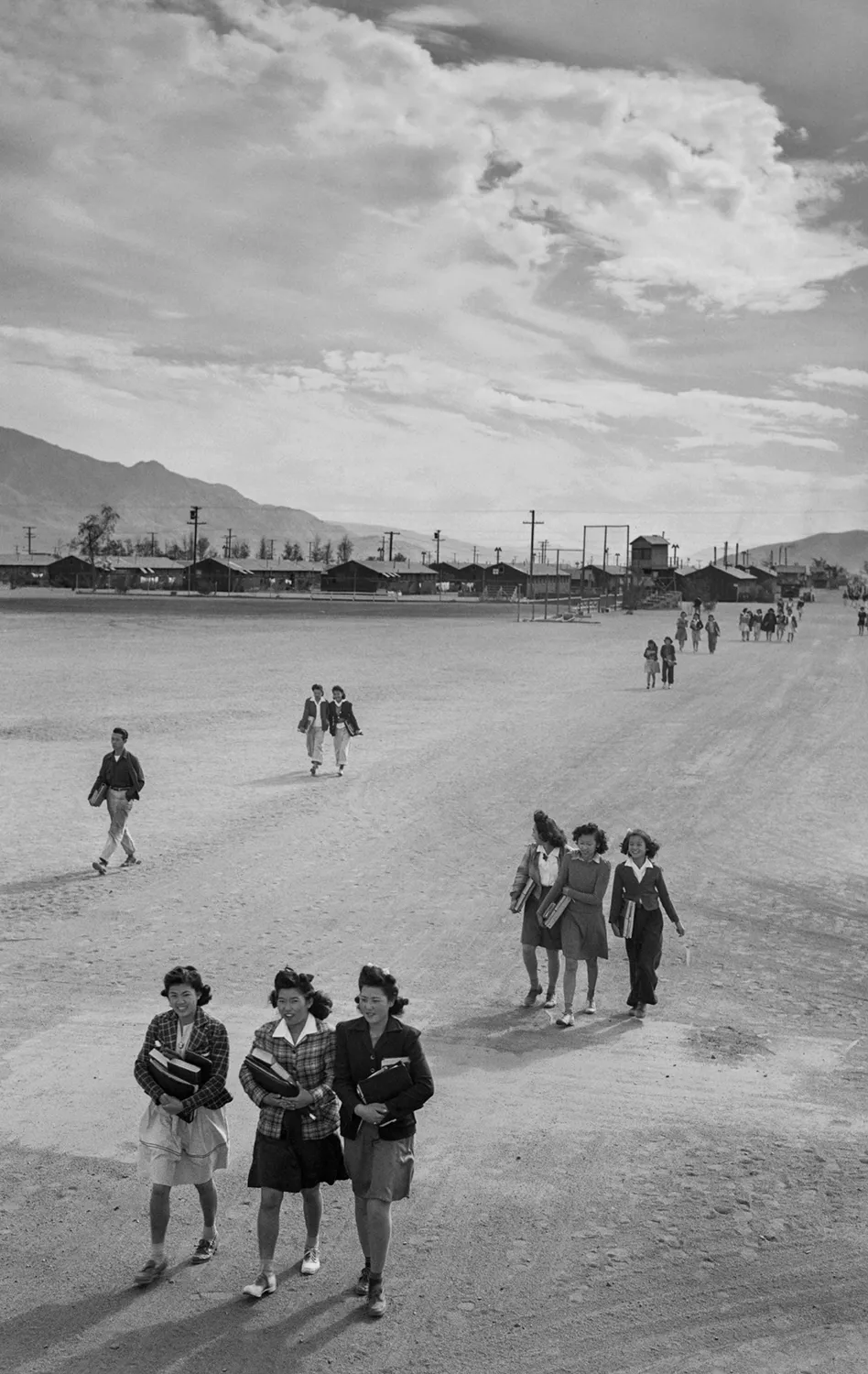Japanese American internees walking across the flat desert landscape of Manzanar camp, with barracks and mountains visible in the distance under a cloudy sky