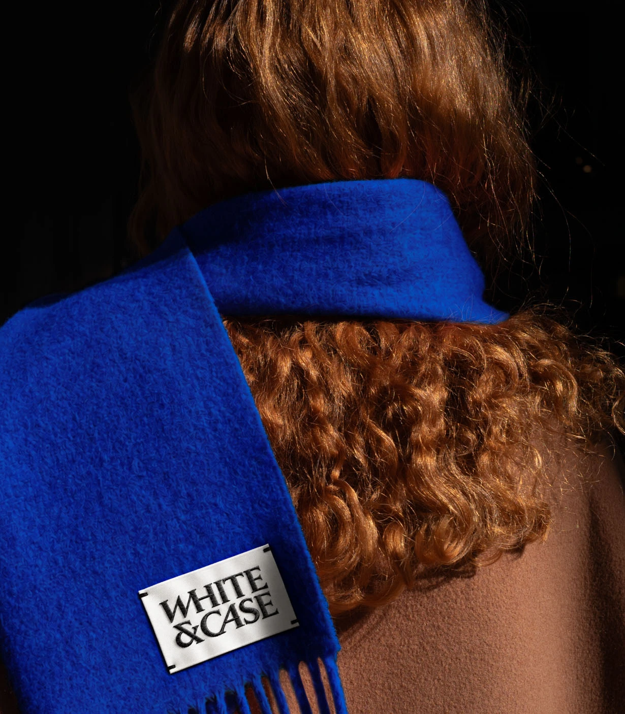 Person wearing a bright blue White & Case scarf with the brand label visible against brown hair, shot from behind against a dark background