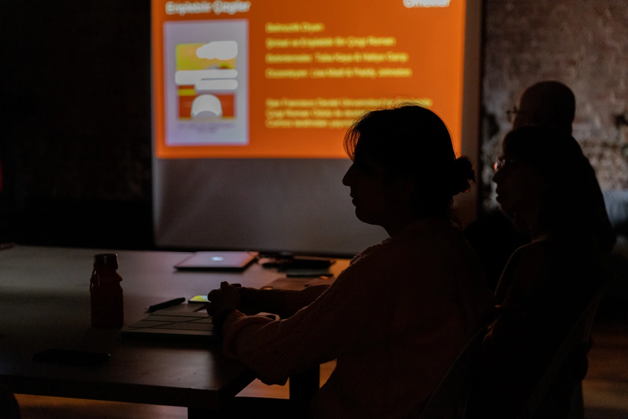 A photo from the Accessible Lines workshop organized in collaboration with British Council Turkey. There is a presentation projected on the wall in a dark environment, the participants sitting at the table are seen from the side.