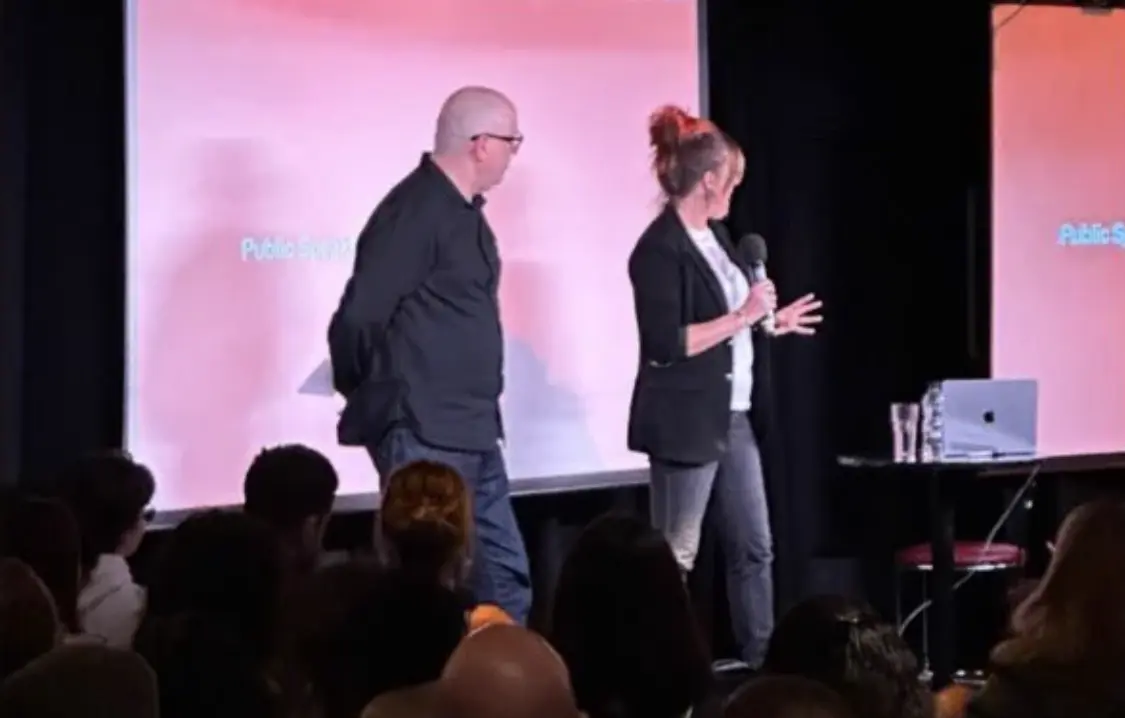 Man in dark jacket and glasses and woman in black blazer speaking on stage at TypoCircle's 50th anniversary event, with pink-lit backdrop and audience in foreground