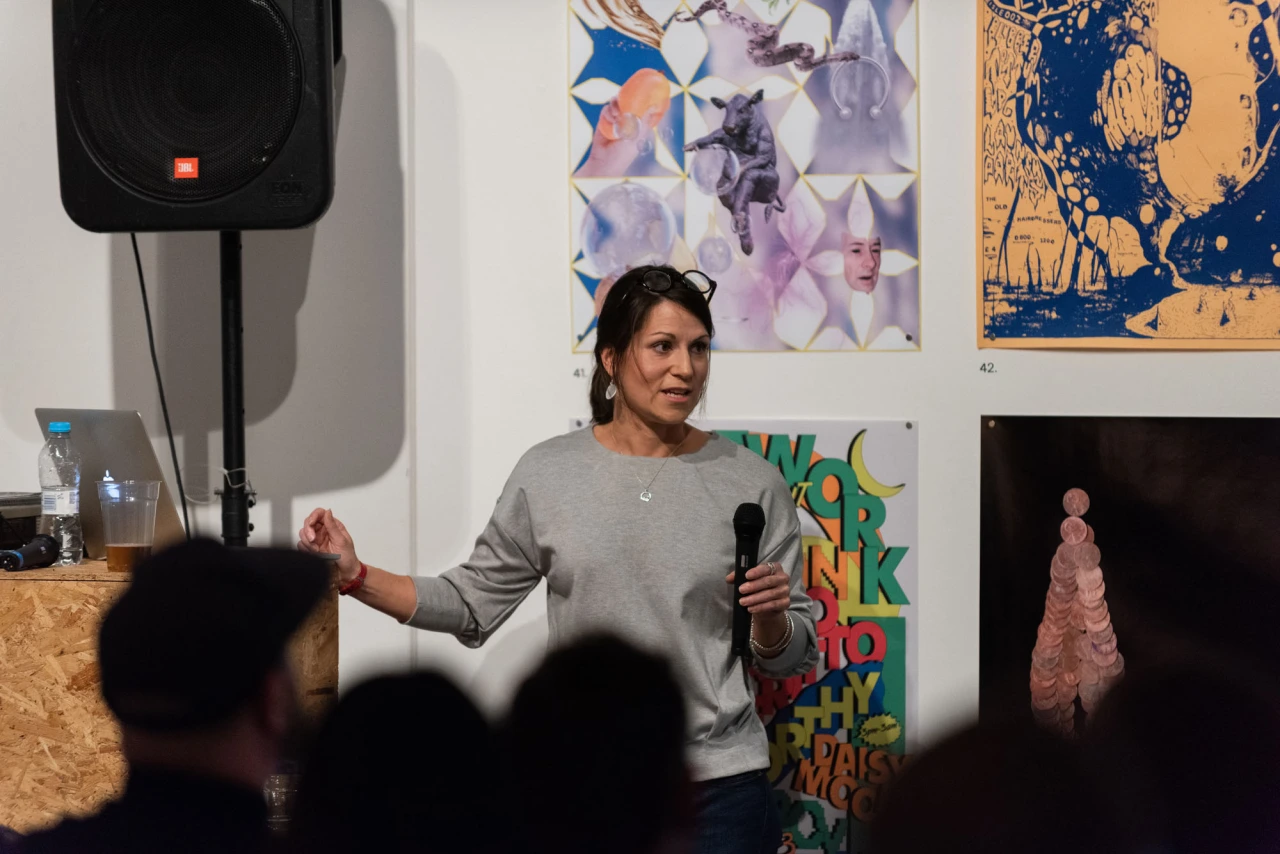 Woman speaking at a podium during a presentation, with colourful artwork displayed on the wall behind her and an audience in the foreground.