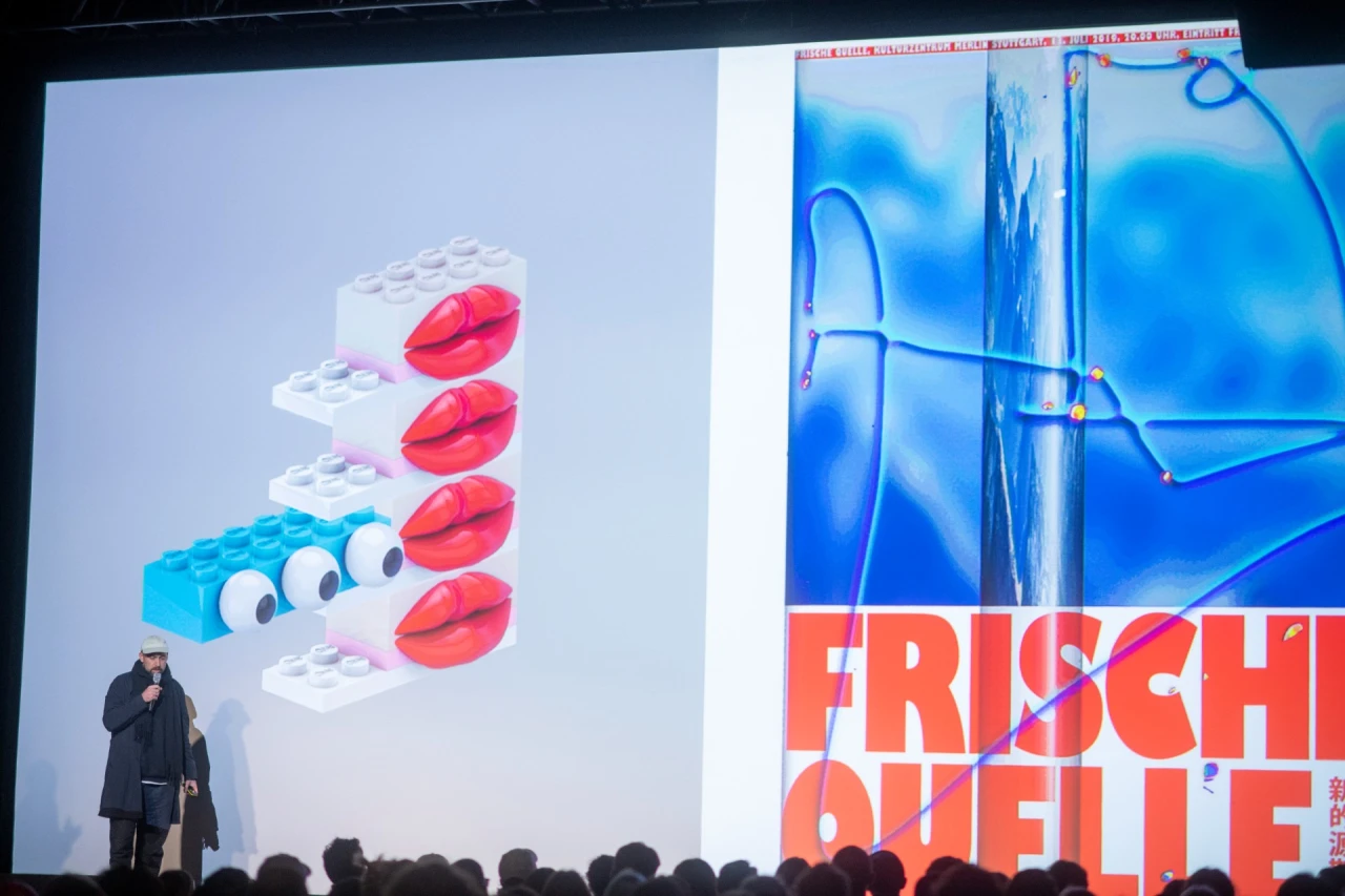 Person standing before large screen displaying stacked white, blue and red plastic bricks with bright pink lips, beside text reading "Frisch Quelle
