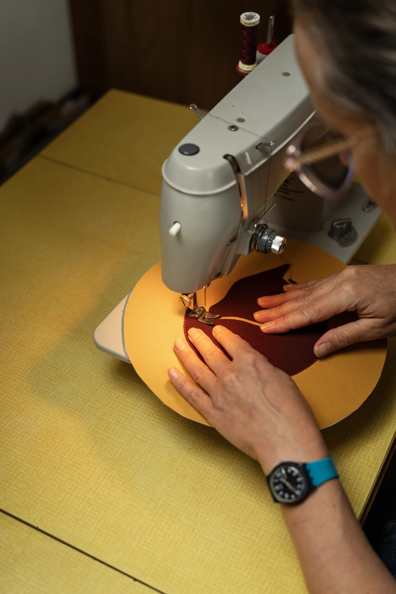 Hands guiding mustard-yellow fabric under a sewing machine needle, stitching burgundy thread onto the material on a yellow work surface