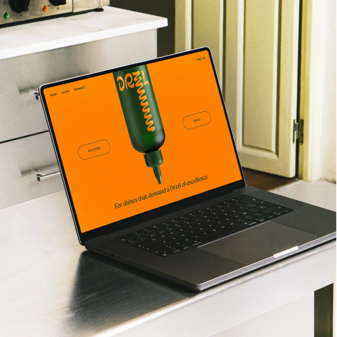 Laptop displaying Drzzl's orange website homepage with a green and gold olive oil bottle illustration on a desk beside kitchen cabinetry