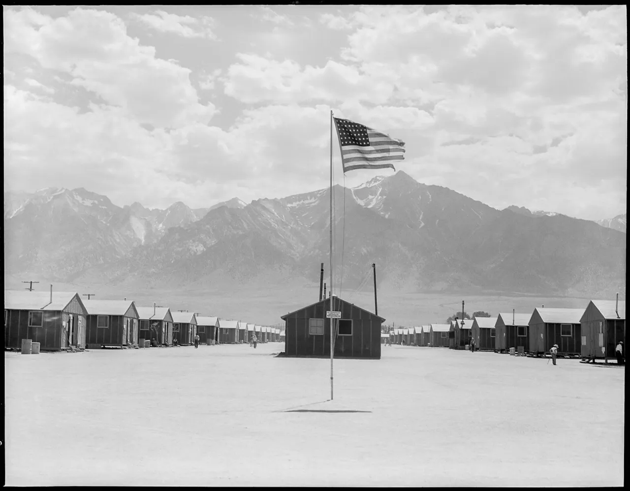 American flag flying above a central building at Manzanar internment camp, with barracks lining both sides and snow-capped mountains in the distance