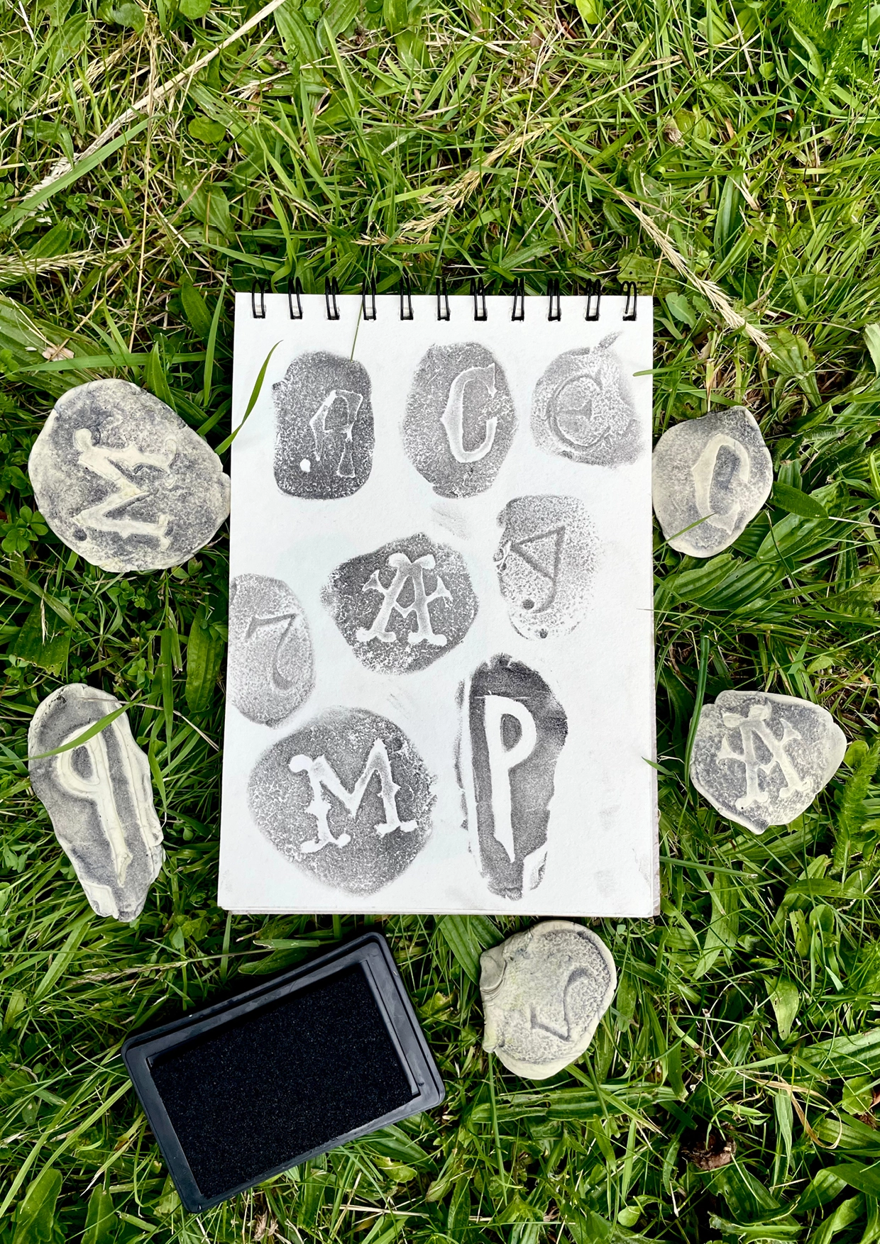 Spiral notebook with hand-printed letterforms surrounded by stone tiles bearing impressed letters and a black ink pad on grass
