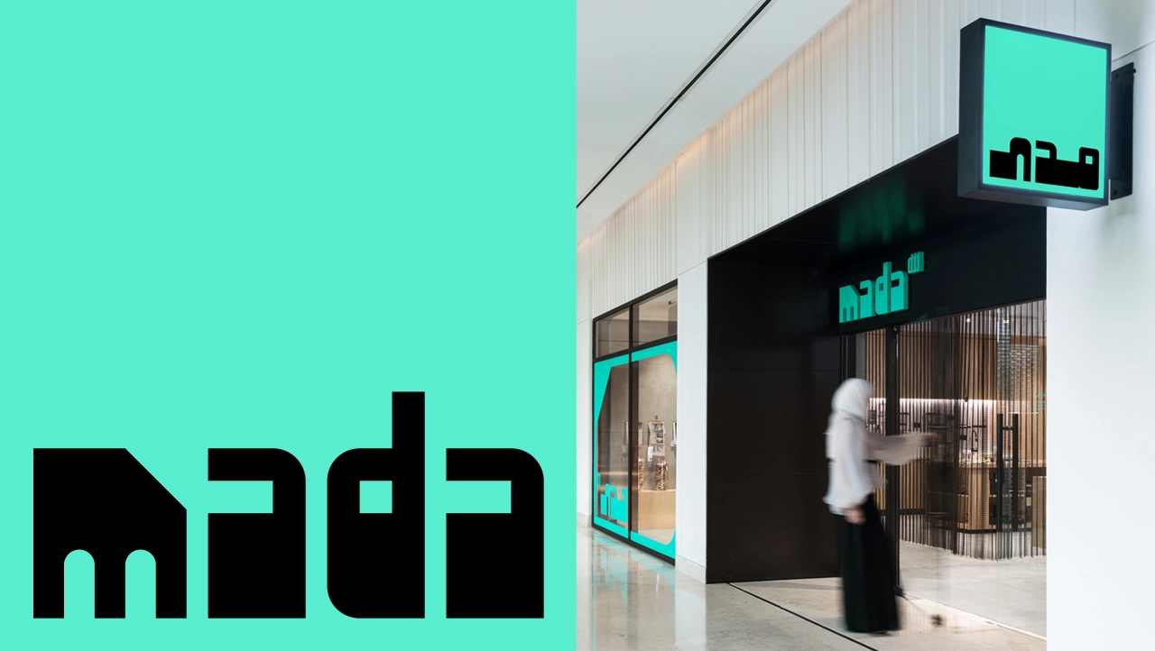 Mada storefront with turquoise signage and logo on white and black facade, with a blurred figure walking past