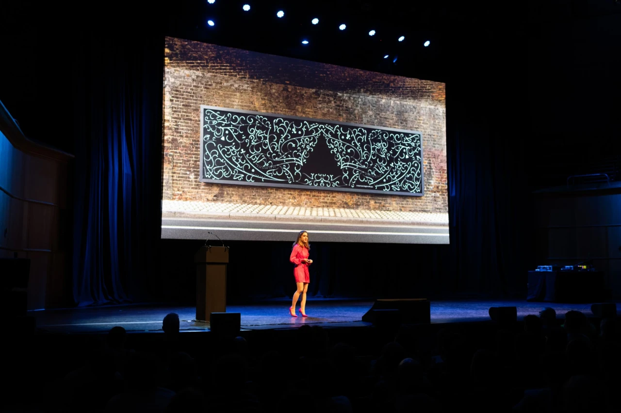 Speaker in red dress on stage presenting to audience with large screen displaying ornamental black and white artwork on brick wall