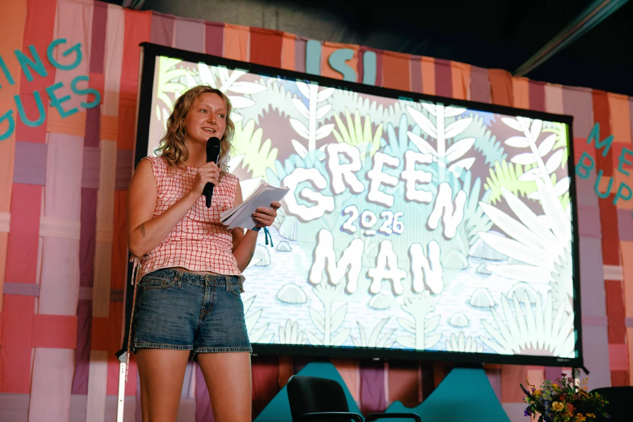A woman in a red-and-white checked top and denim shorts speaks into a microphone on stage in front of a backdrop displaying "Green Man 2026."