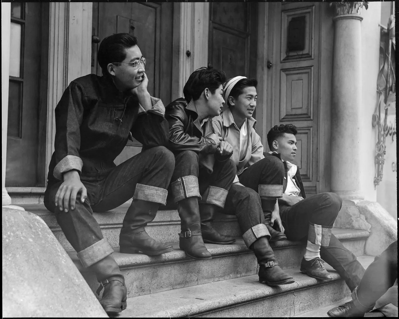 Four men and boys sit together on steps outside a wooden building at Manzanar internment camp, smiling and looking off to the side in this black and white photograph