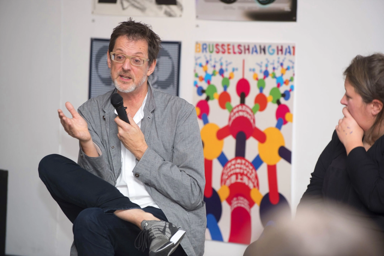Man in grey shirt gestures while speaking at International Assembly design festival event, with colourful poster visible on wall behind him