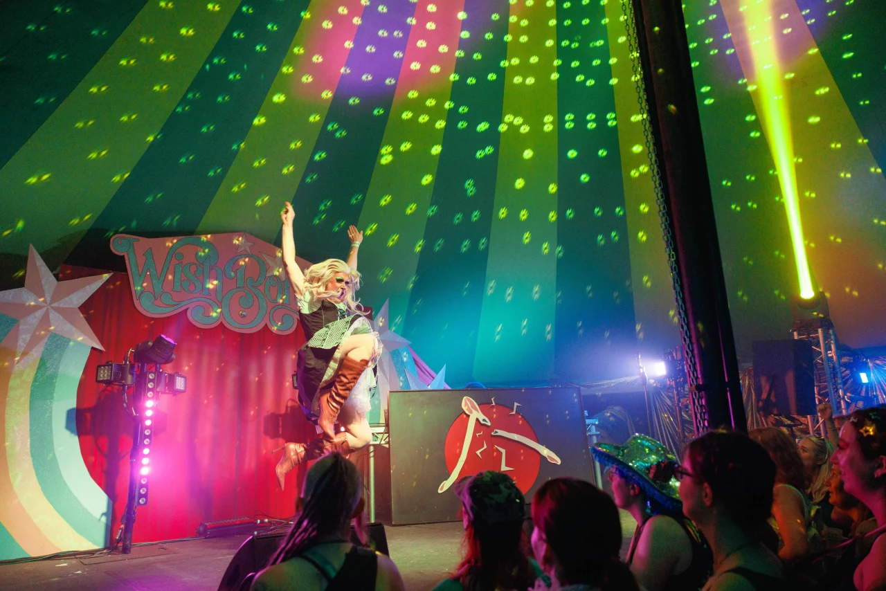 A performer in a dress and boots leaps energetically into the air during a colorful, tented stage performance.