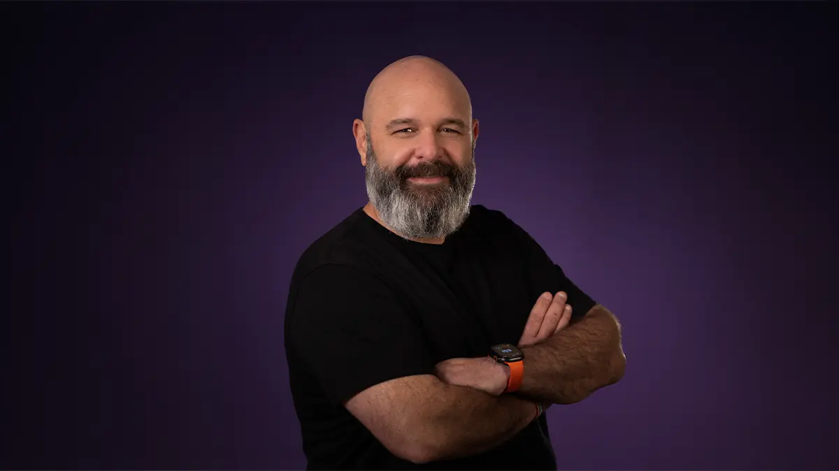 Bald man with grey beard wearing a black t-shirt, smiling with arms crossed against a purple background