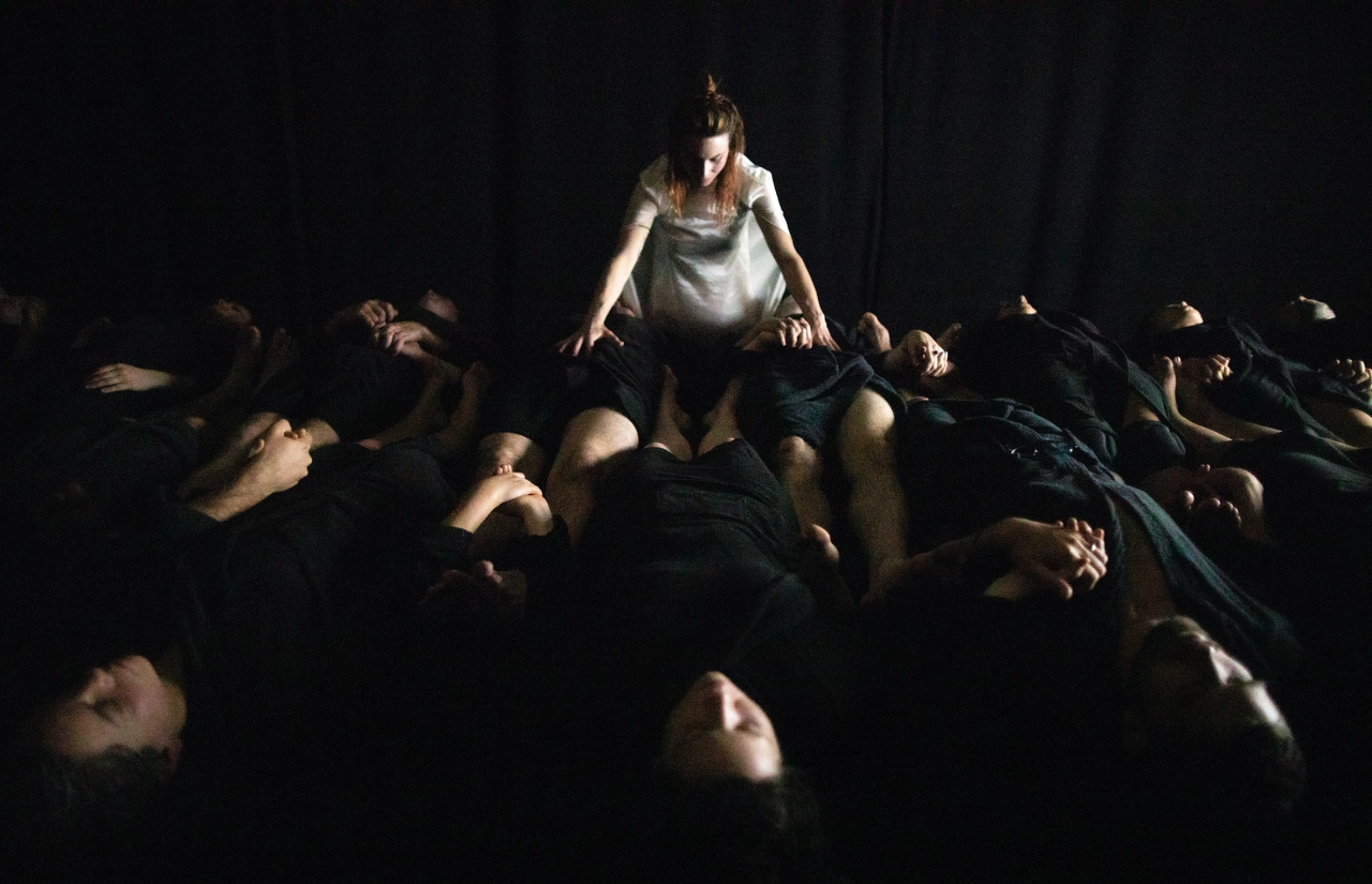 A dancer in pale clothing stands among prone participants lying on the ground during an intense immersive performance in dim lighting
