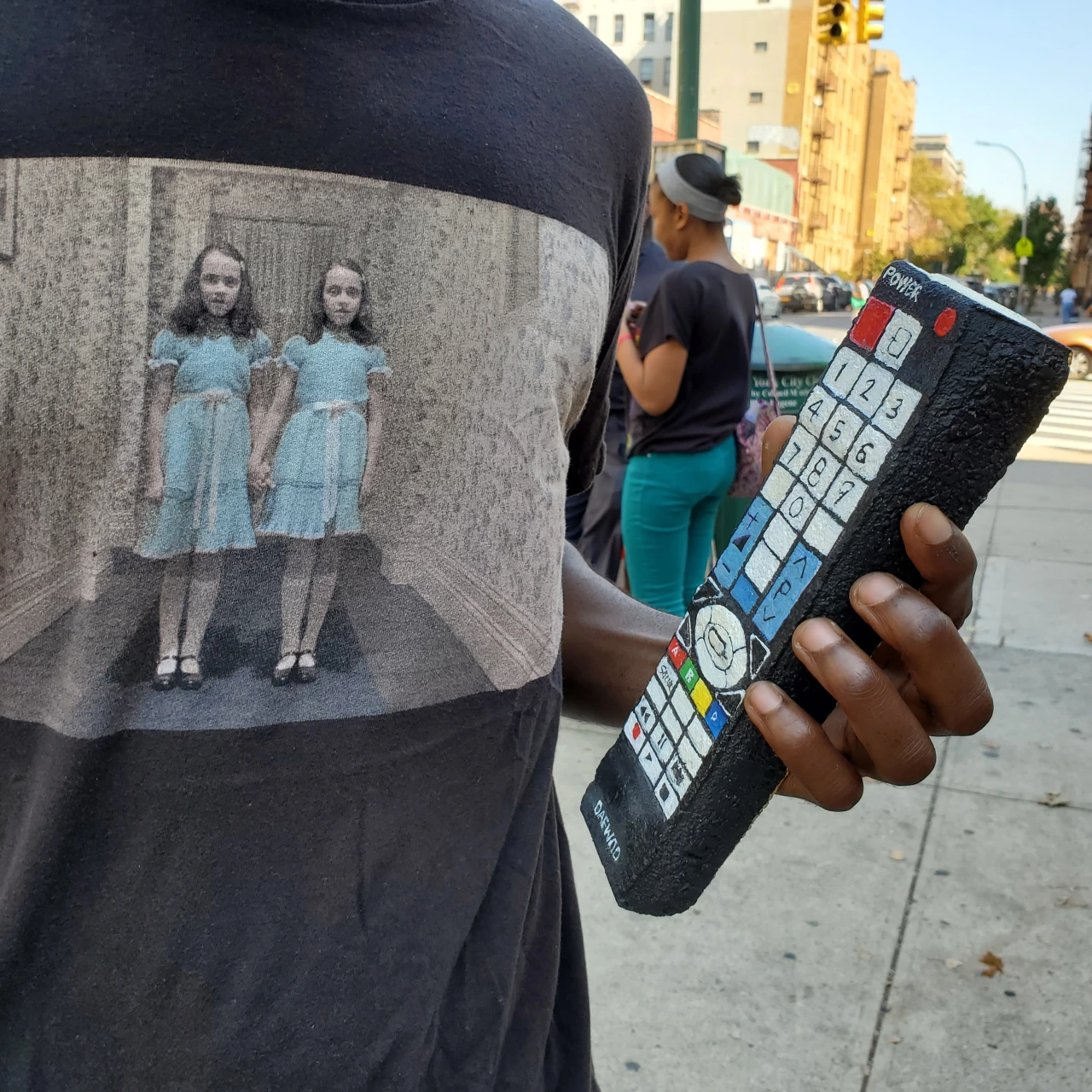 Person wearing a t-shirt printed with two girls in blue dresses holds a painted rock depicting a mobile phone keypad on an urban street