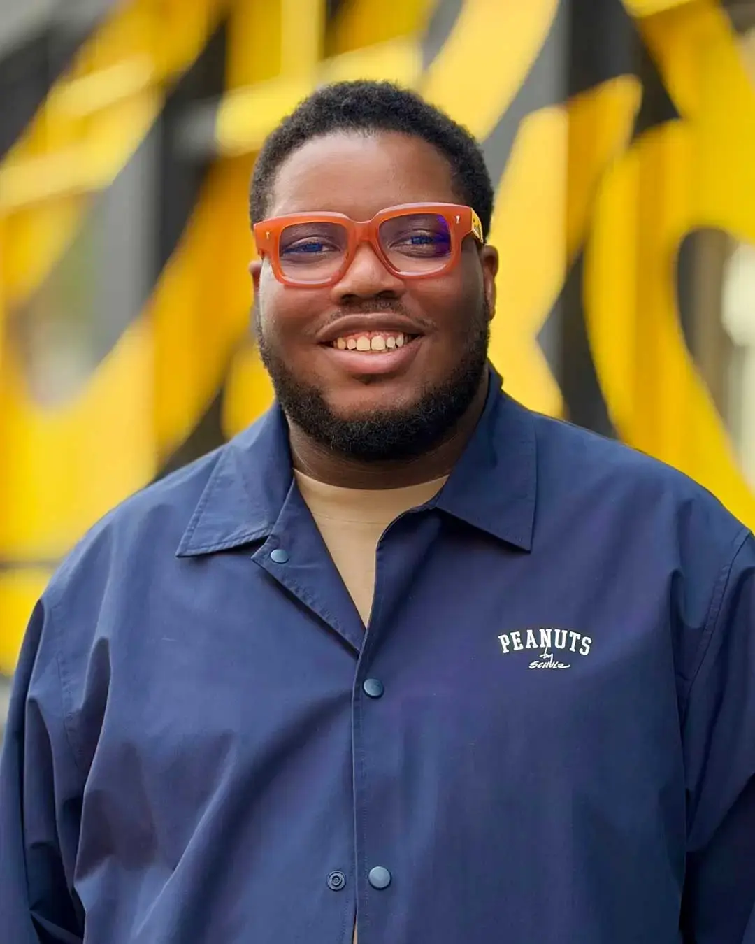 Lánre Adeleye smiling at the camera wearing orange-framed glasses and a navy Peanuts Snoopy shirt, with yellow brushstroke shapes blurred in the background