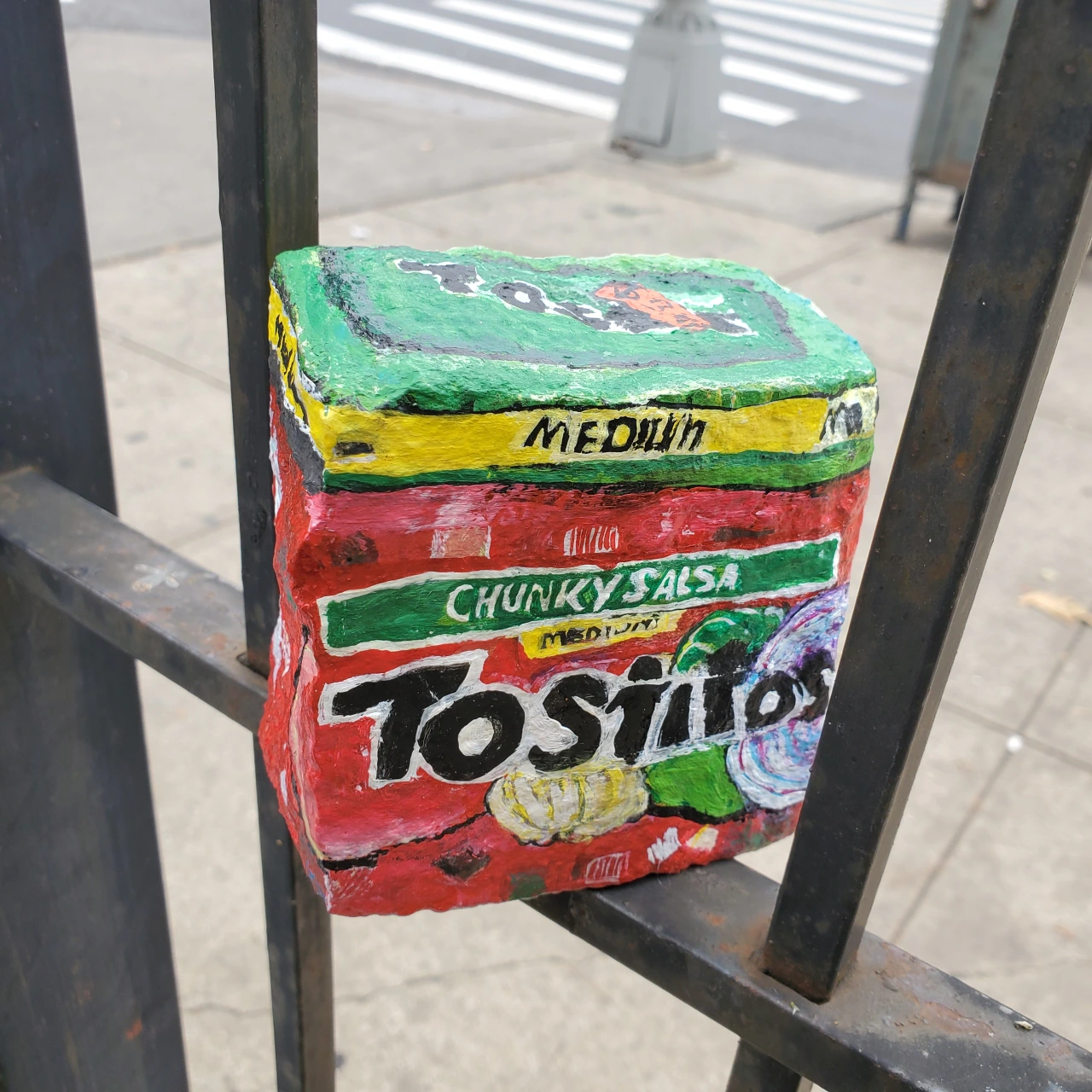 Painted rock depicting a Tostitos Chunky Salsa jar with green lid and red packaging, propped against a metal railing outdoors