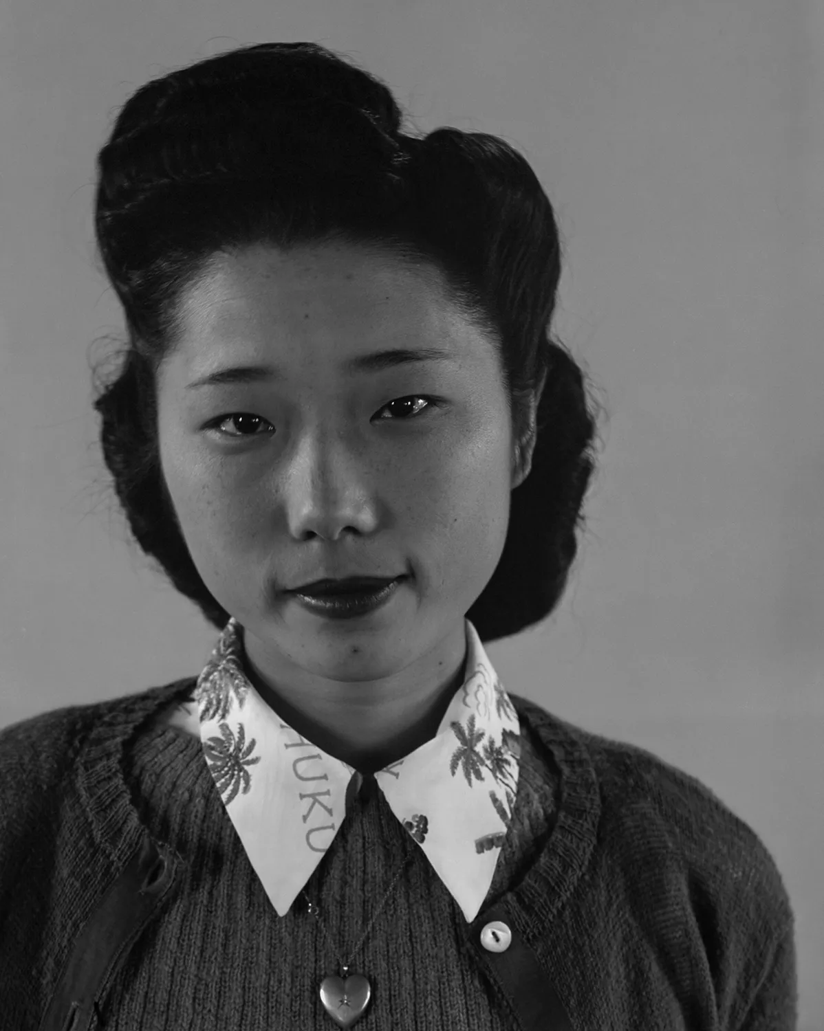 Black and white portrait photograph of a Japanese American woman with dark hair styled in victory rolls, wearing a patterned collar and dark jacket, taken at Manzanar internment camp in the 1940s