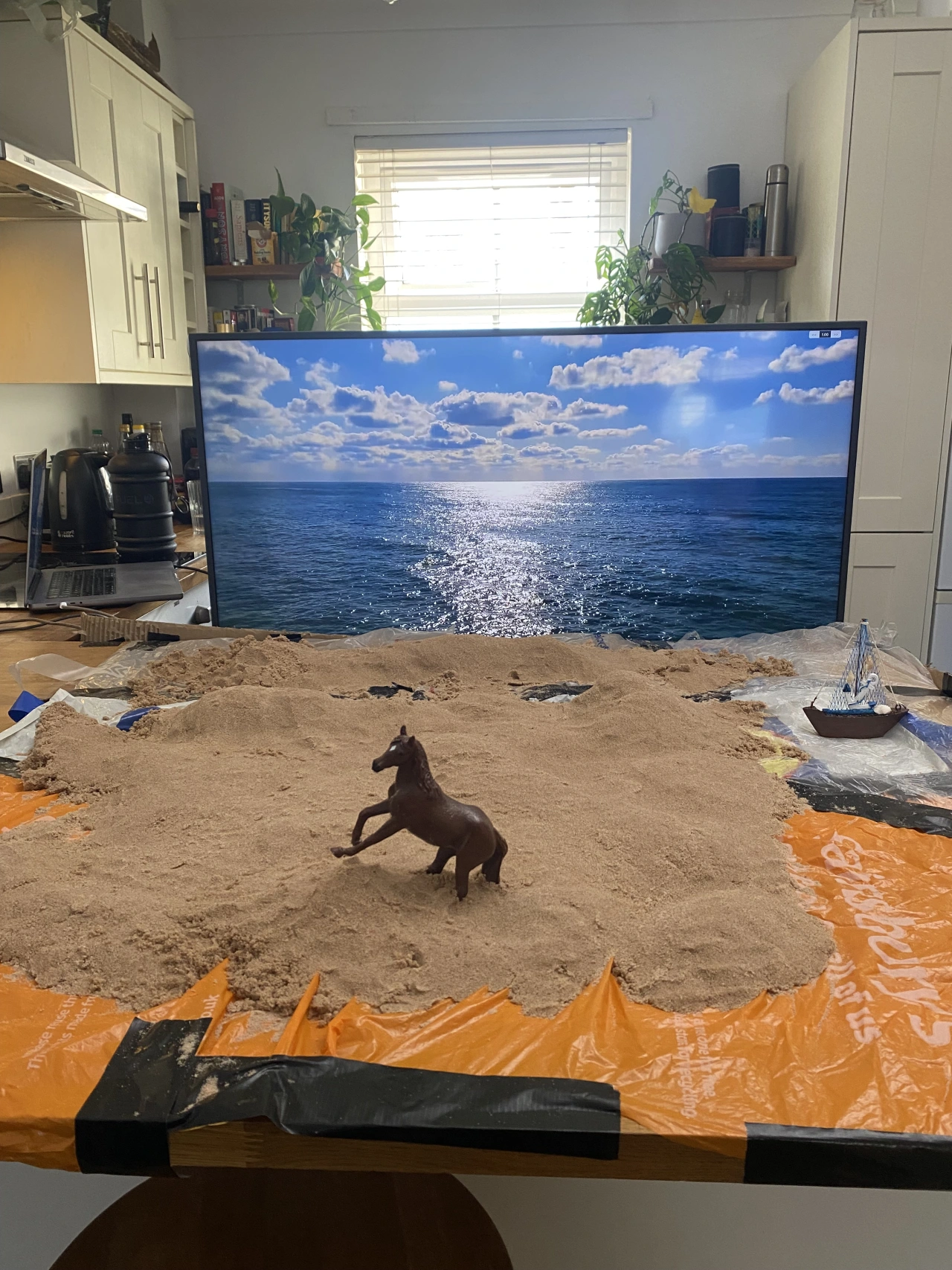 A dark horse figurine stands on sand in front of a monitor displaying an ocean and sky seascape in a domestic kitchen setting