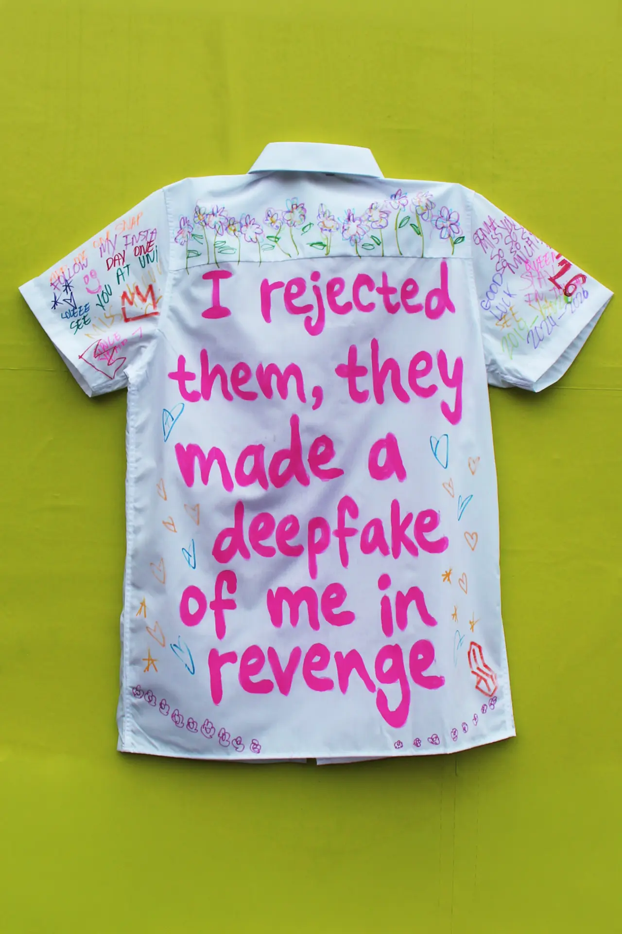 White school leavers' shirt with handwritten messages and drawings, featuring large pink text reading "I rejected them, they made a deepfake of me in revenge