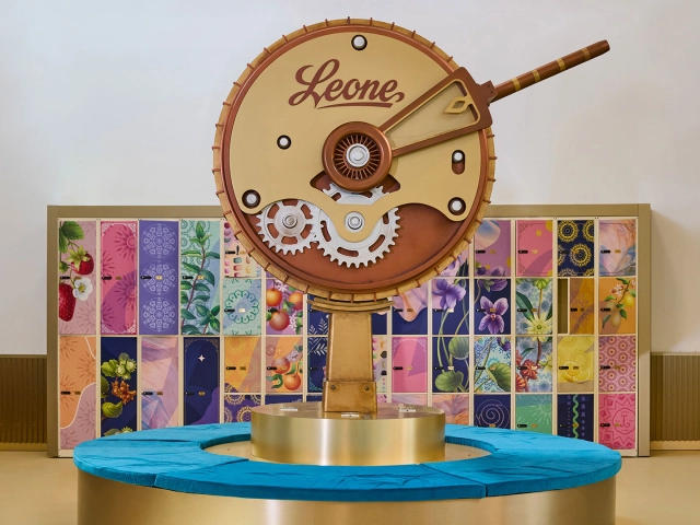 Leone's giant wooden clock face with exposed gears mounted above a blue circular platform, with colourful packaged sweets displayed on shelving behind it inside the Happiness Factory in Turin