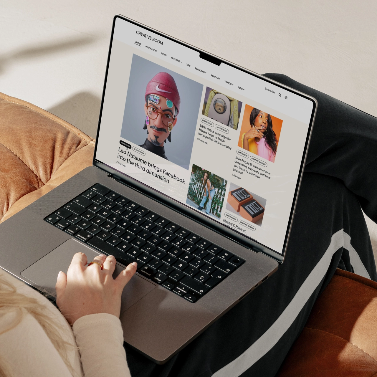 A laptop displaying the Creative Boom website with various article thumbnails and images, held by someone's hand resting on the keyboard
