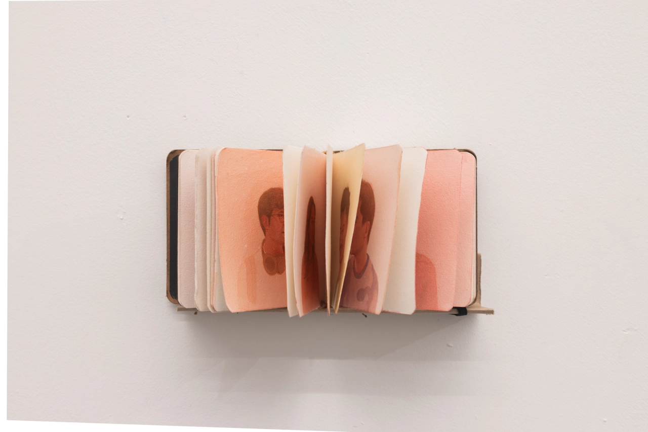 An open accordion-style booklet displaying fanned pages with abstract portraits in warm peachy and terracotta tones against a white wall