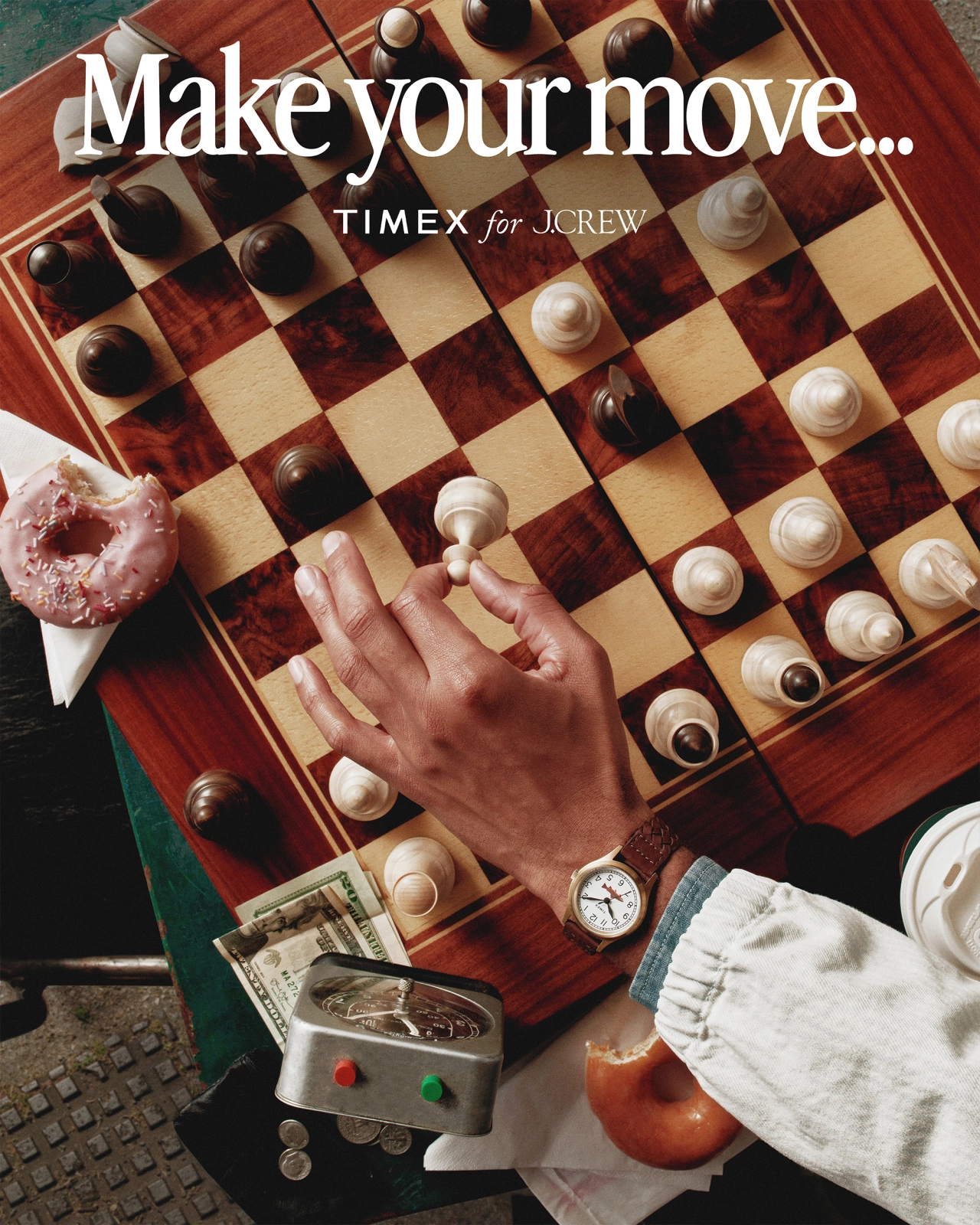 A person's hand hovers over a chess board with white and cream pieces, wearing a Timex watch, surrounded by a doughnut, onion and coins in a styled still life advertisement for Timex for J.Crew