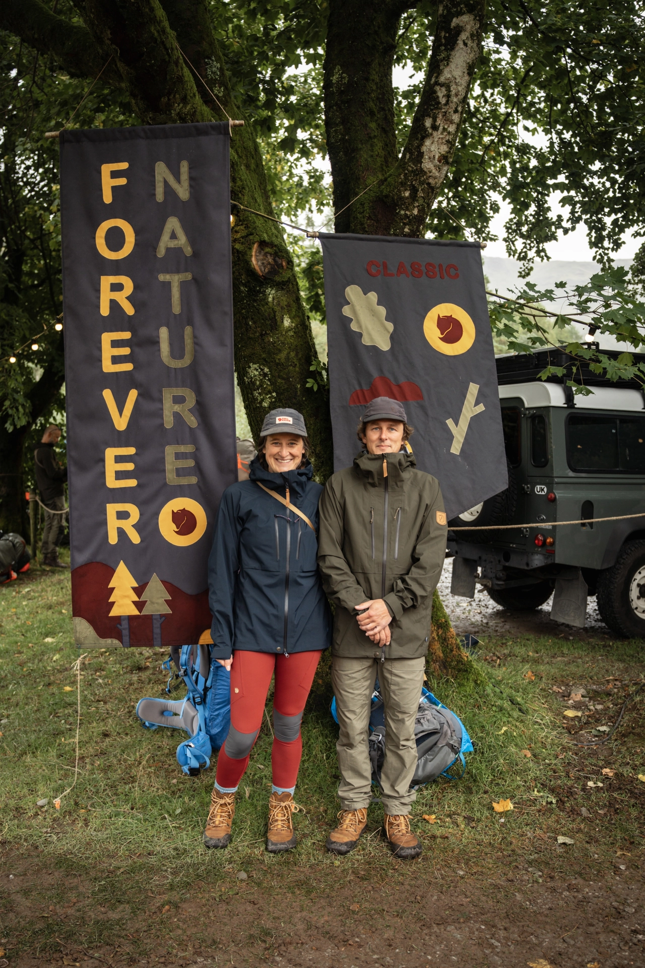 Emma and Andrew Groves stand in front of two large hand-stitched banners displaying Fjällräven branding, with a vehicle and moss-covered trees behind them