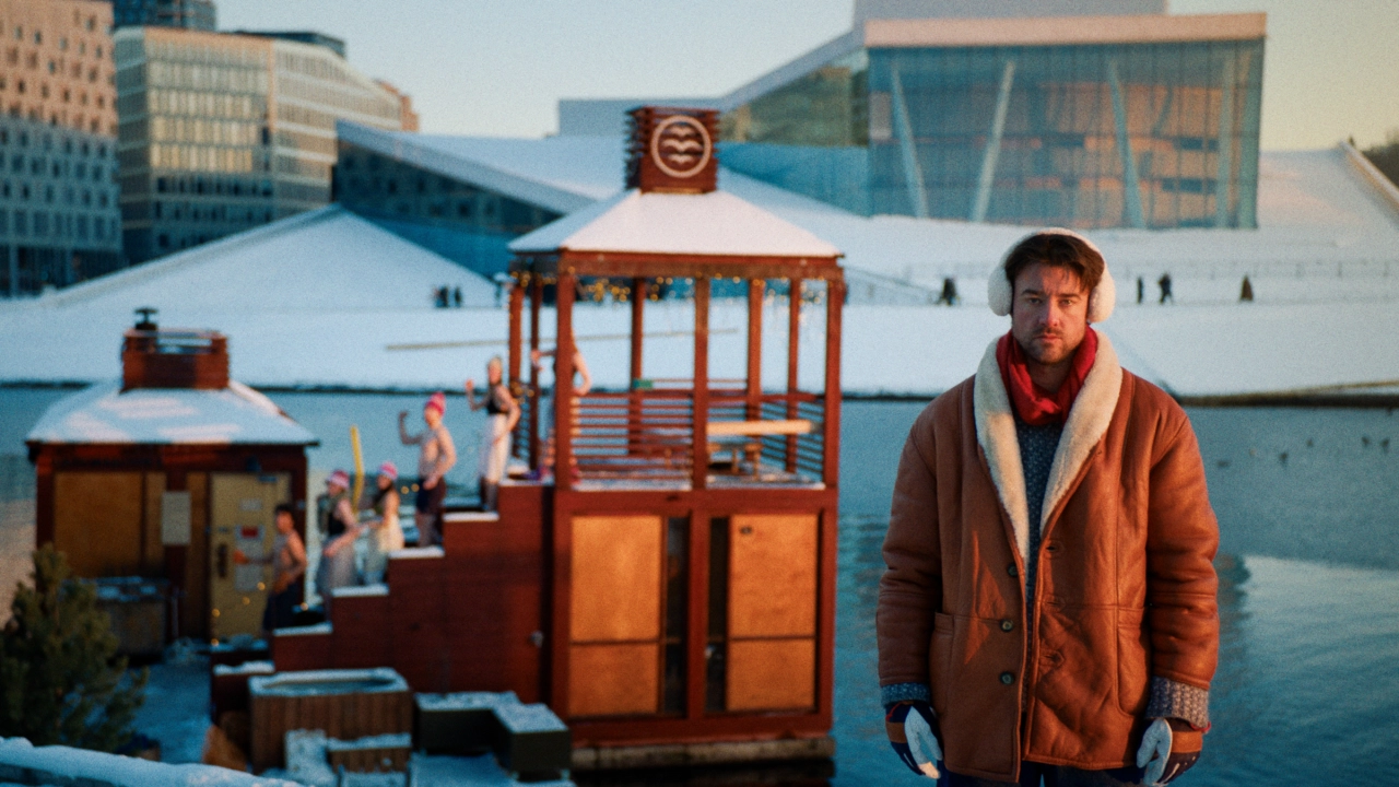 Man in rust-coloured jacket stands on frozen Oslo harbour with colourful wooden structures and snow-covered buildings behind him