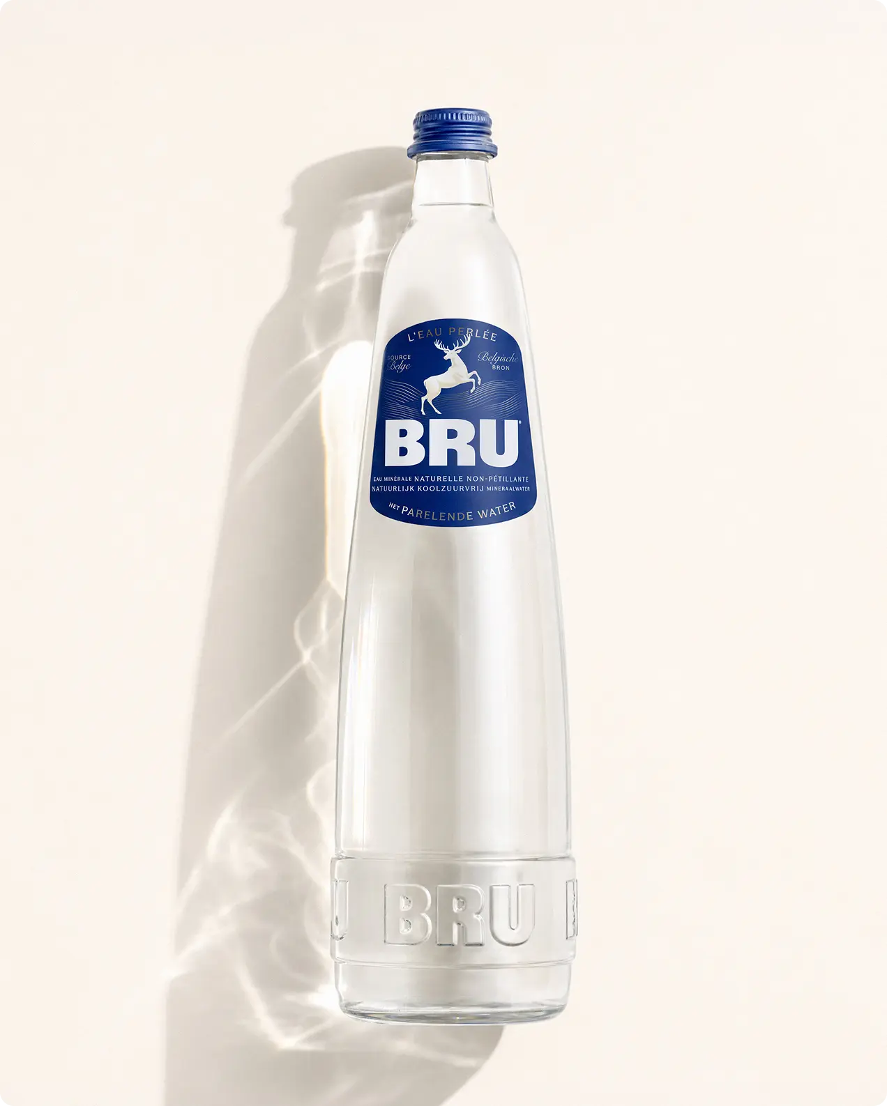 Clear glass bottle of BRU sparkling water with blue label featuring a deer motif and blue screw cap, casting a soft shadow against a cream background