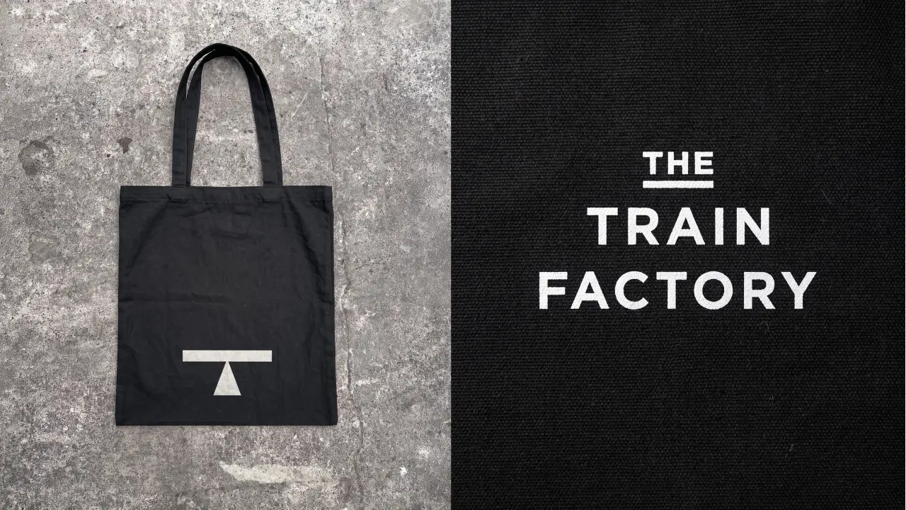 Black tote bag with white minimalist logo featuring a T-shaped lifting mark, displayed against concrete background next to The Train Factory branding