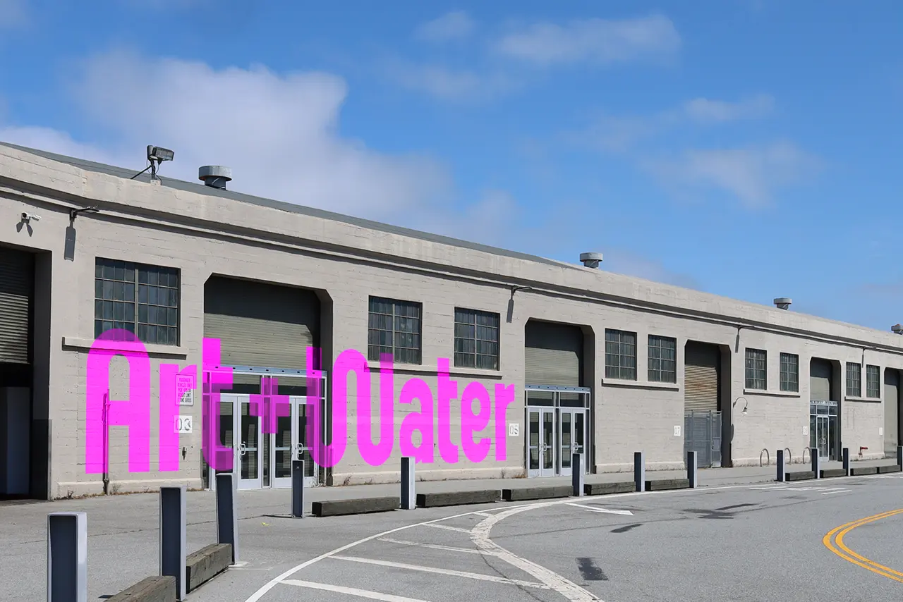 Magenta letters spelling "ART+WATER" on the facade of a modernist waterfront building with large windows and bollards in front