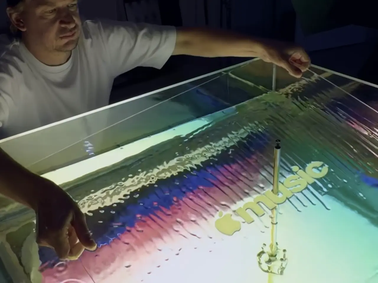 A person gestures over a shallow tray containing mineral oil with colourful sound wave patterns and a musical note created by vibrations from a transducer
