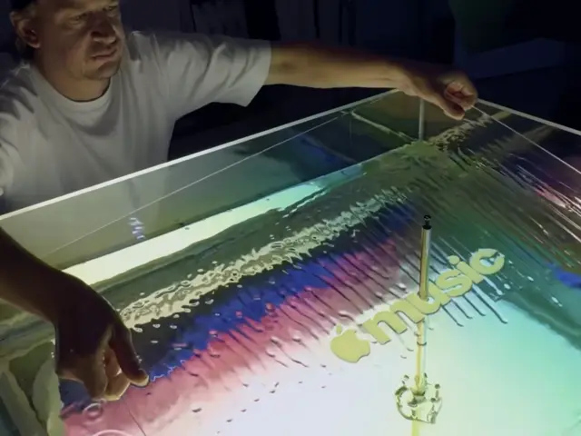 A person gestures over a shallow tray containing mineral oil with colourful sound wave patterns and a musical note created by vibrations from a transducer