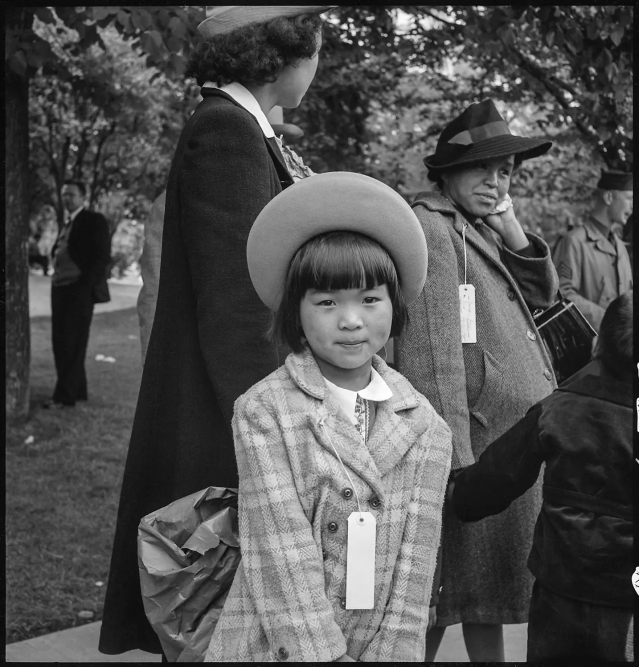 Young Japanese American girl wearing a wide-brimmed hat and coat with identification tag, standing with adults at Manzanar internment camp