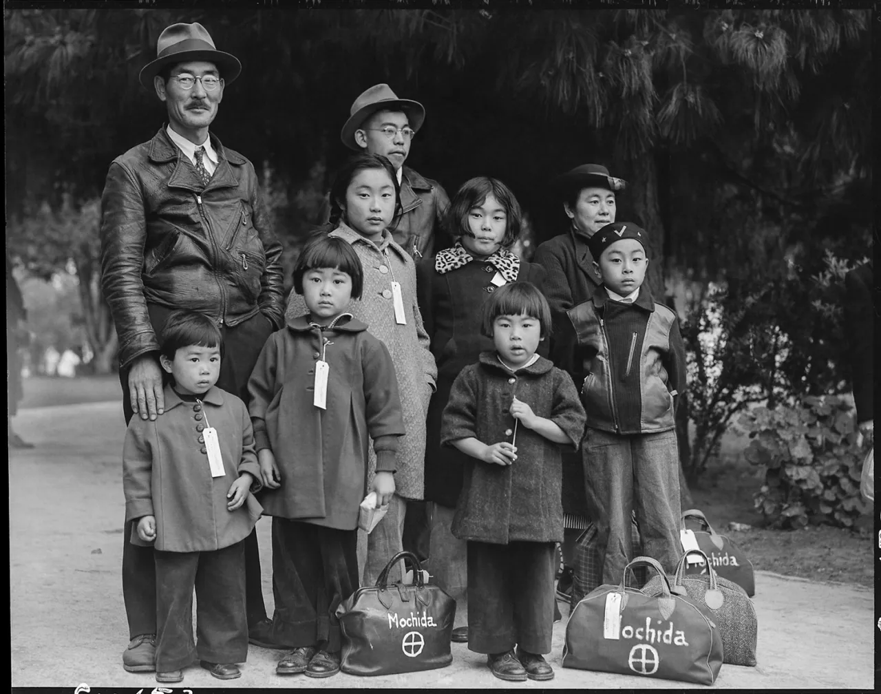 Japanese American family with luggage labelled "Inochida" stands together at Manzanar internment camp in the 1940s
