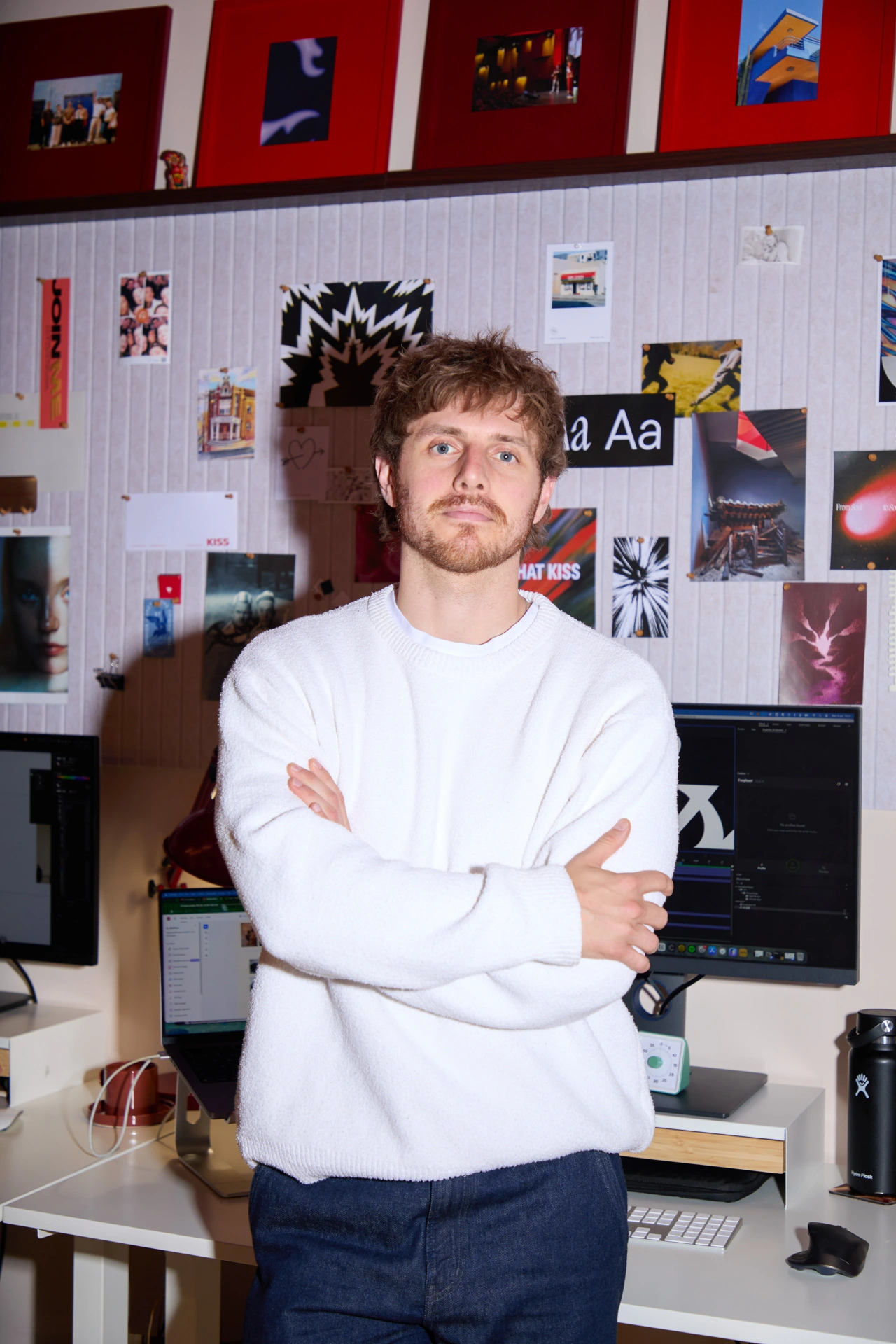 Man with crossed arms standing at desk in creative studio with mood board of images and designs on wall behind him