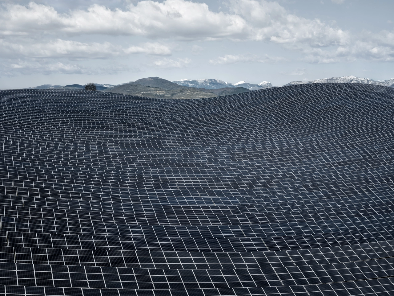 Vast expanse of solar panels stretching across rolling terrain towards snow-capped mountains under a cloudy sky
