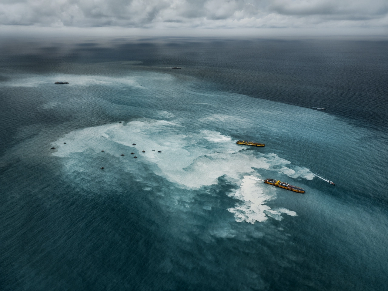 Aerial view of a shallow turquoise lagoon with white sandy bottom surrounded by darker ocean waters, with two yellow and red cargo ships positioned nearby
