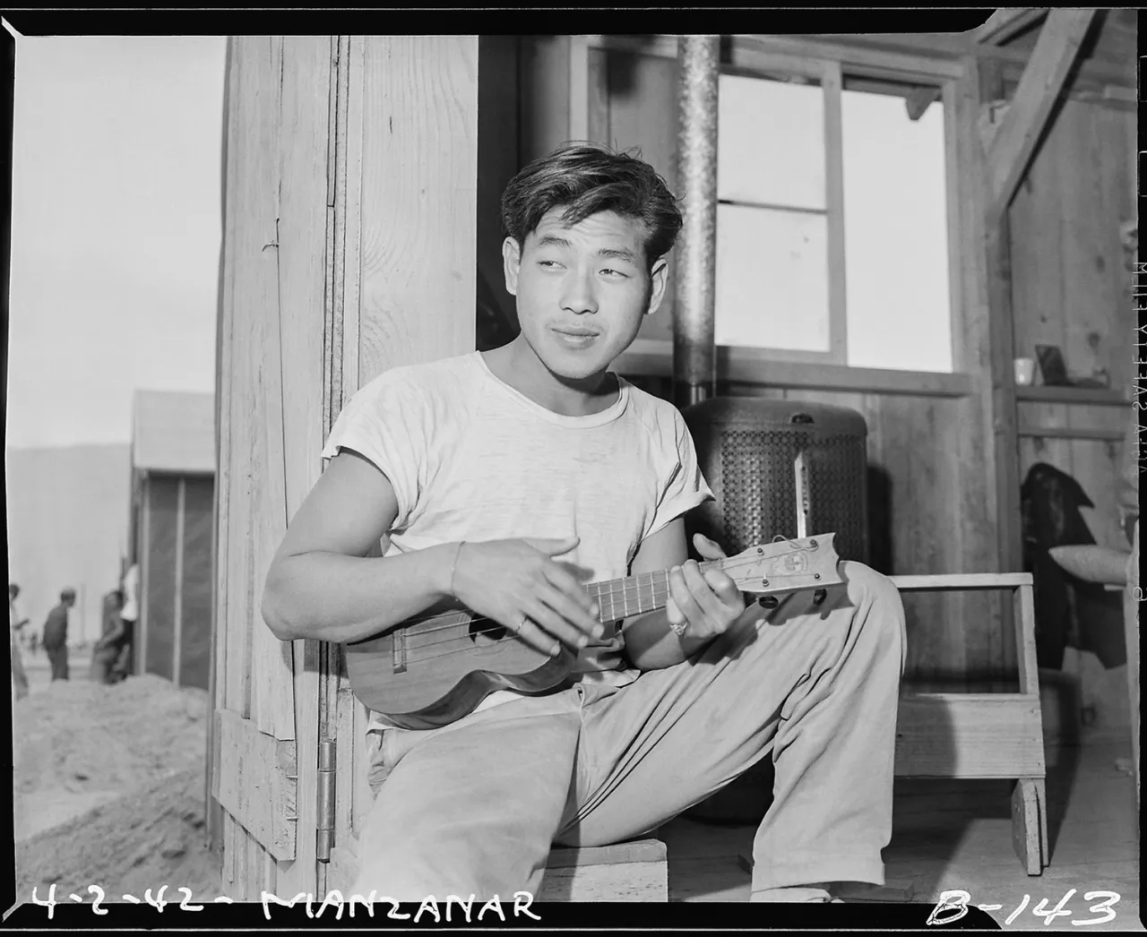 Man playing guitar while seated against a wooden post inside a Manzanar barracks, 1942