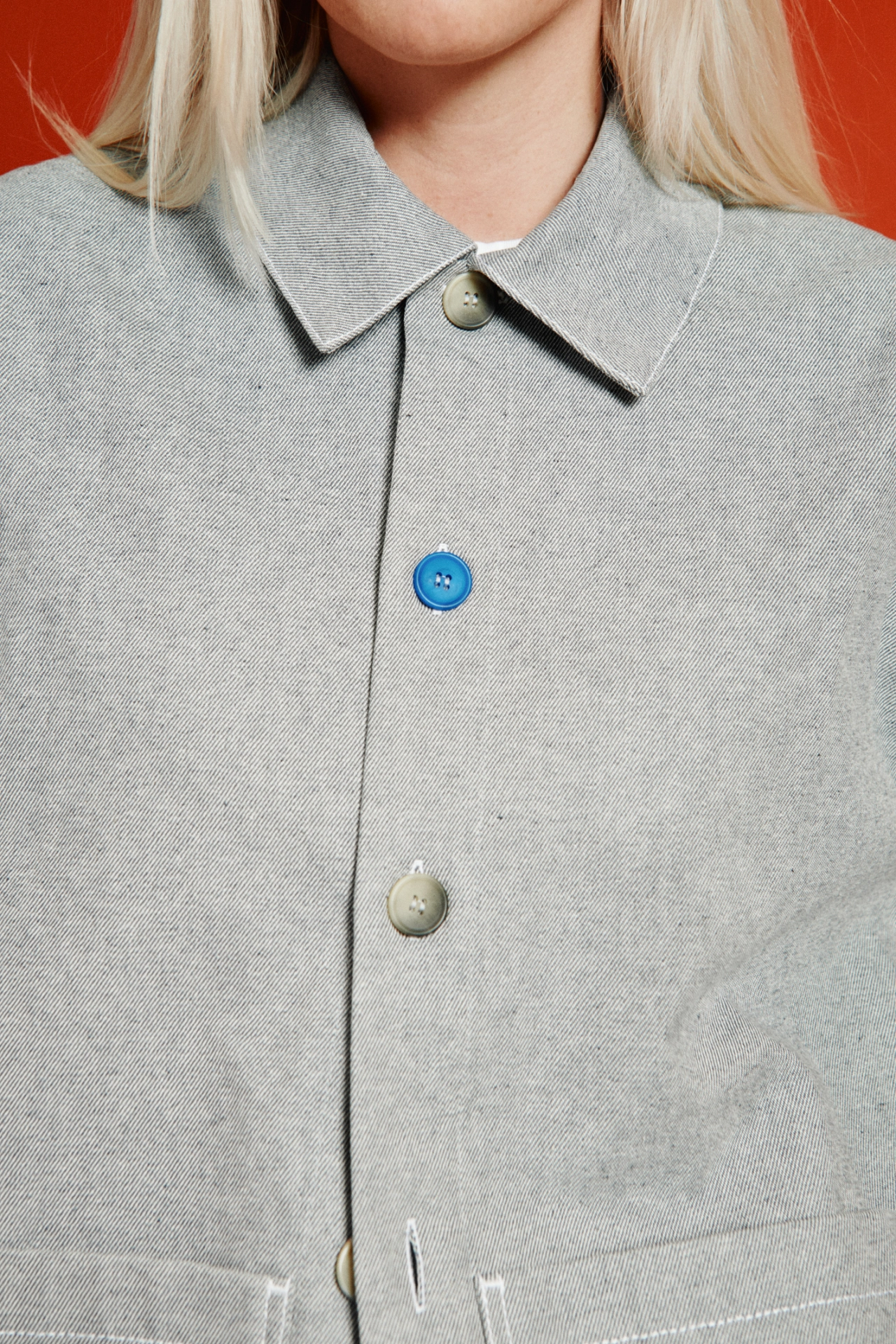 Close-up of a light grey shirt with a pointed collar, featuring a bright blue button alongside neutral-toned buttons, worn against a red background