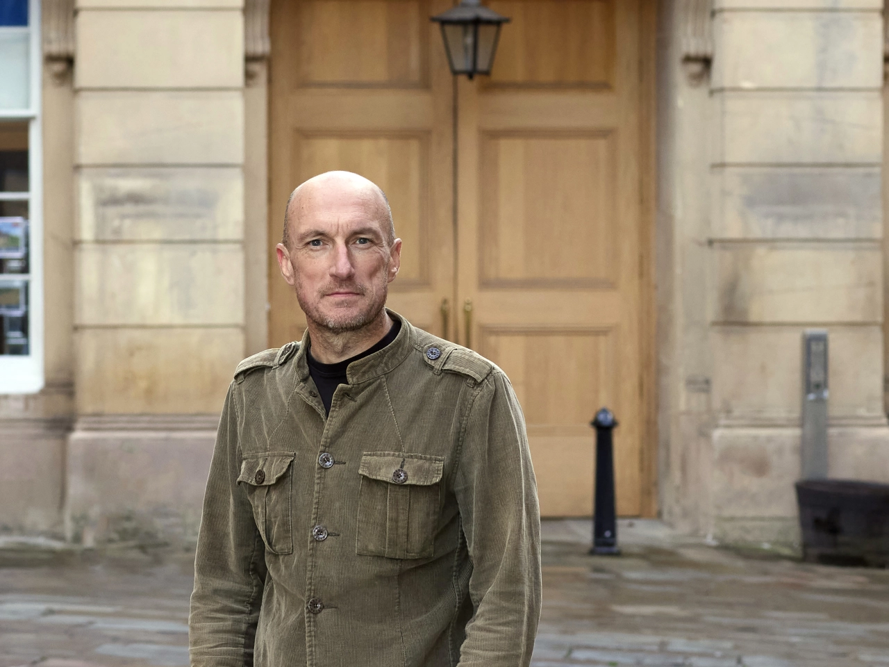 Tony Clarkson stands in front of a large wooden door on a stone building, wearing a khaki shirt jacket