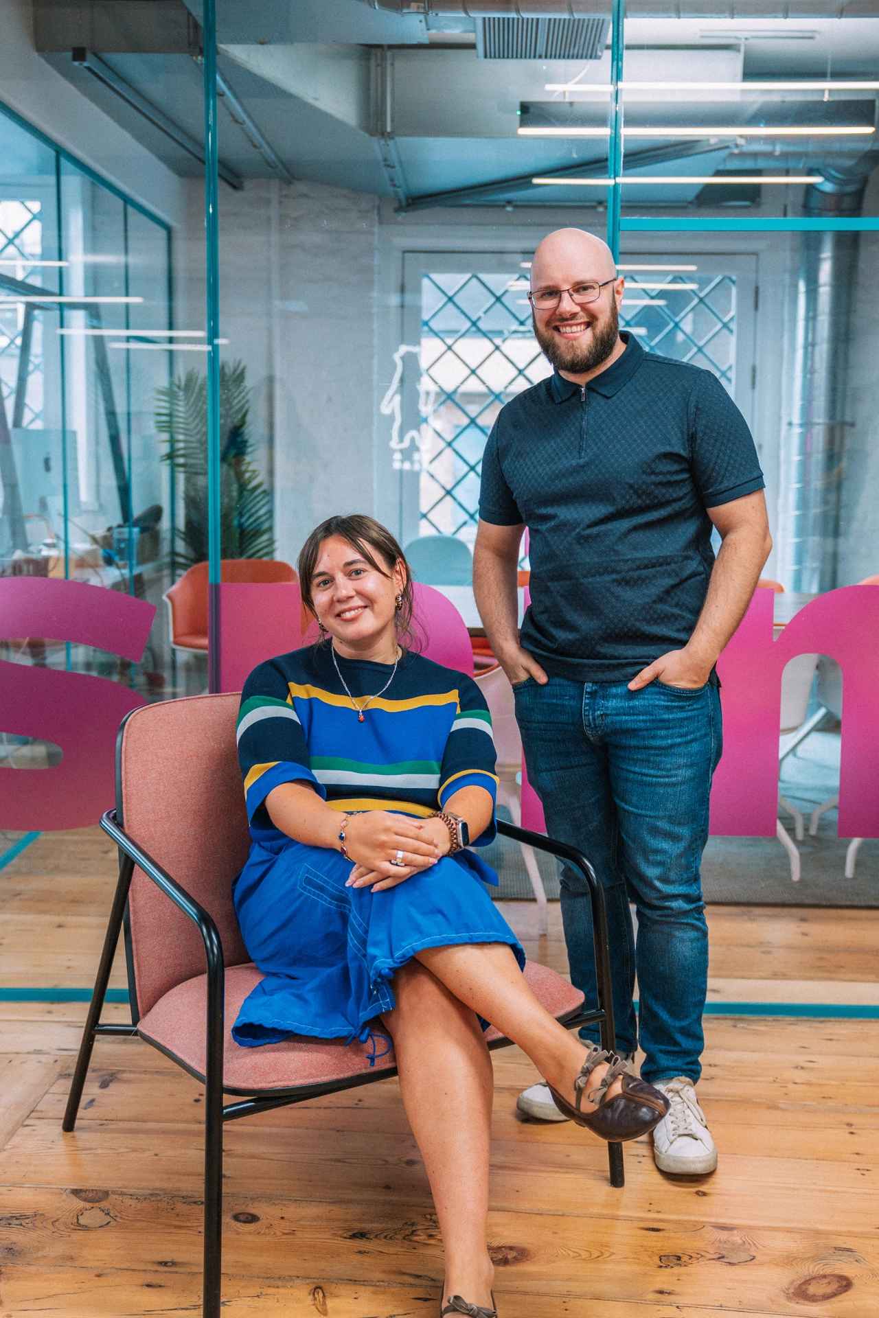 Woman seated on pink chair wearing striped jumper and blue skirt beside standing man in dark polo shirt in modern office with glass partitions and turquoise structural beams