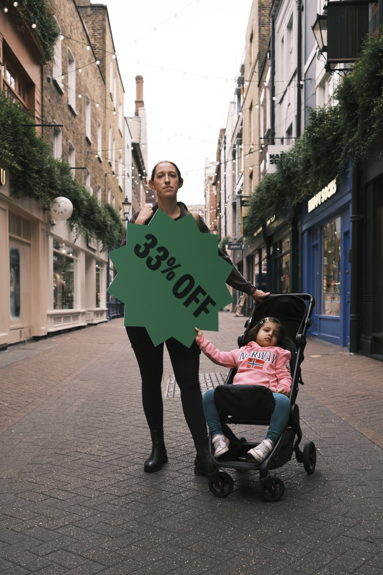 Woman holding a large green starburst sign reading "33% off" while standing with a child in a pram on a city street